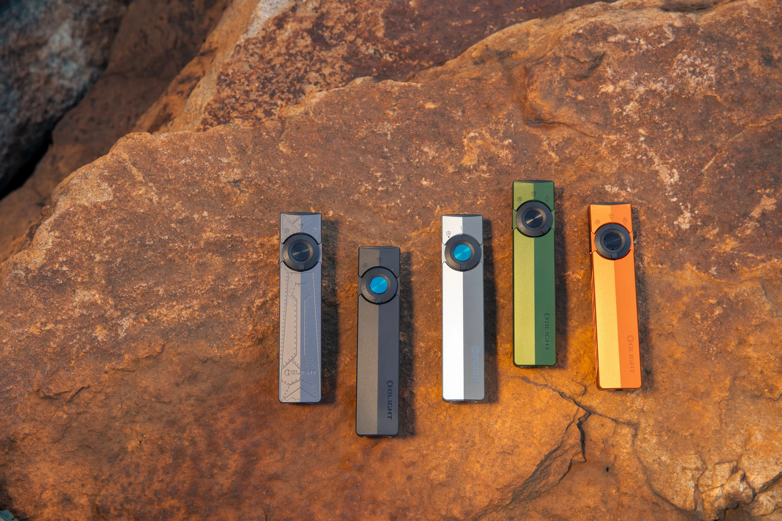 Cinco linternas Olight de colores (gris, verde, naranja) sobre roca marrón.